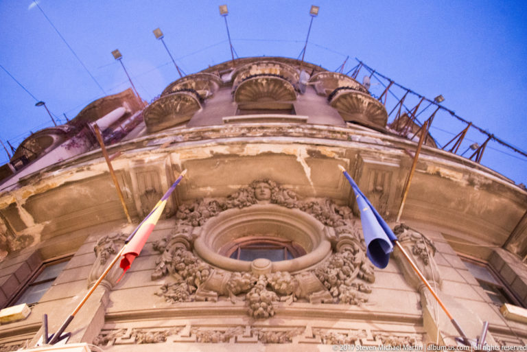 Low-angle view of an ornate oval window with carved scrolls, floral ornament, masks, and two projecting flags.