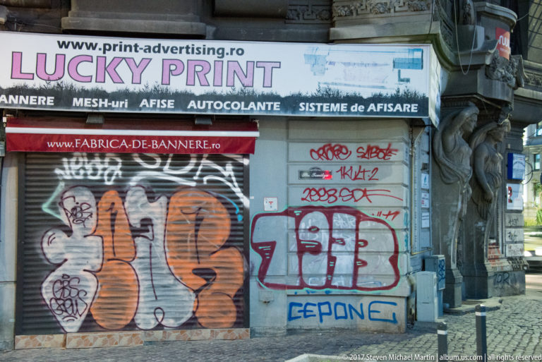 Closed Bucharest storefront with graffiti-covered shutter and Romanian signs advertising printing, banners, stickers, and display systems.