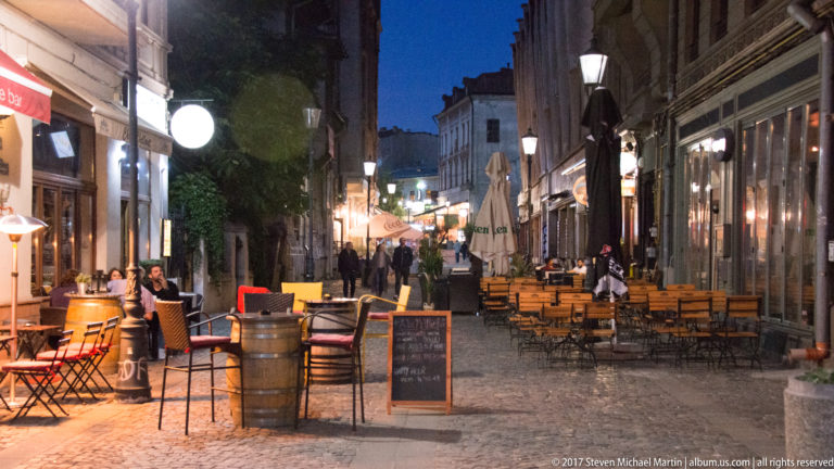Narrow pedestrian street in Bucharest with outdoor restaurant tables, awnings, menu boards, pedestrians, and warm evening lights.