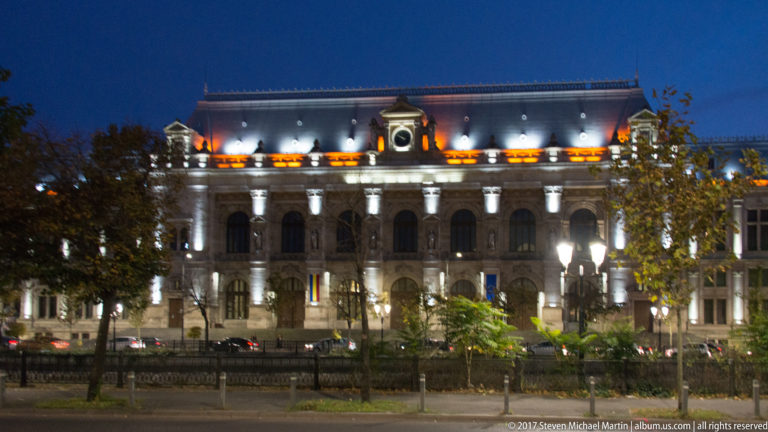 Floodlit historic Bucharest building with arched windows, mansard roof, dormers, Romanian flags, trees, and streetlamps.
