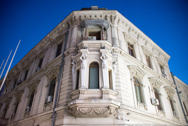White historic Bucharest corner building with projecting bay, carved brackets, pilasters, arched windows, and molded stone ornament.