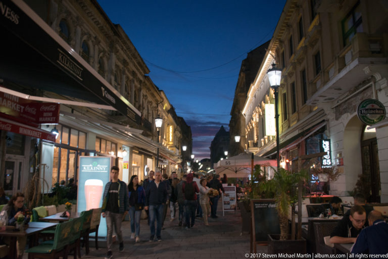 Crowded Bucharest pedestrian street with bars, cafés, outdoor seating, illuminated signs, historic façades, and evening pedestrians.