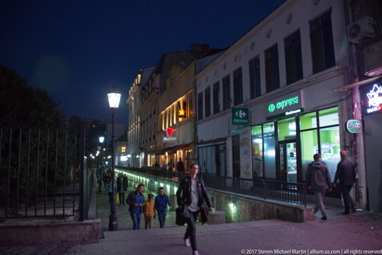 Sunken Bucharest pedestrian walkway beside shops, railings, streetlamps, and people descending through the evening commercial district.