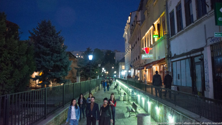Lower-level Bucharest pedestrian corridor with glowing retaining walls, railings, storefronts above, and groups walking toward the camera.