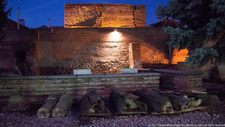 Night-lit archaeological site in Bucharest with brick foundations, masonry walls, gravel, and rows of carved stone fragments.