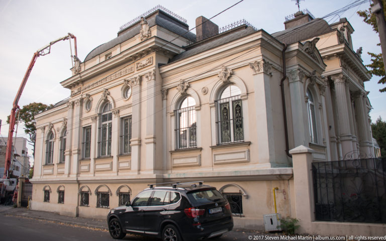 Pale historic Bucharest building with arched windows, pilasters, carved trim, raised basement, and a dark SUV at the curb.