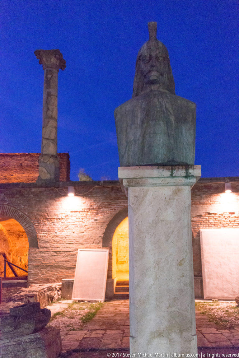 Dark sculpted bust on a pale stone pedestal amid brick ruins, arched openings, and interpretive panels at night.
