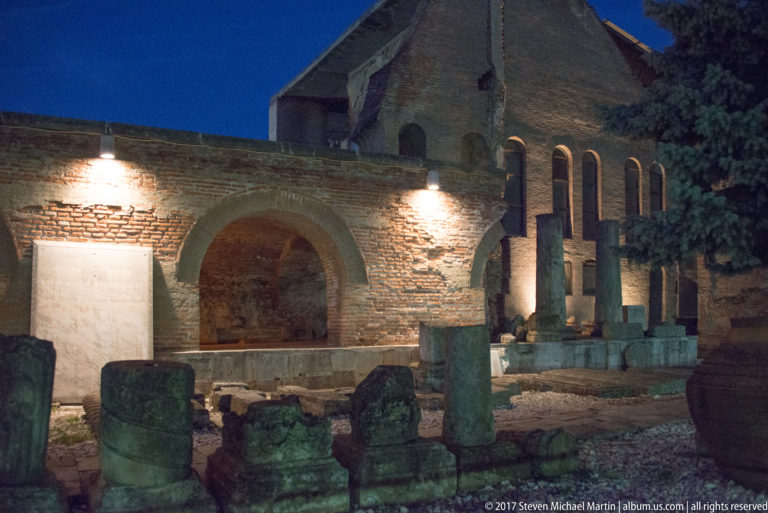 Brick arches, tall openings, stone columns, bases, and carved fragments arranged within an illuminated Bucharest archaeological site.