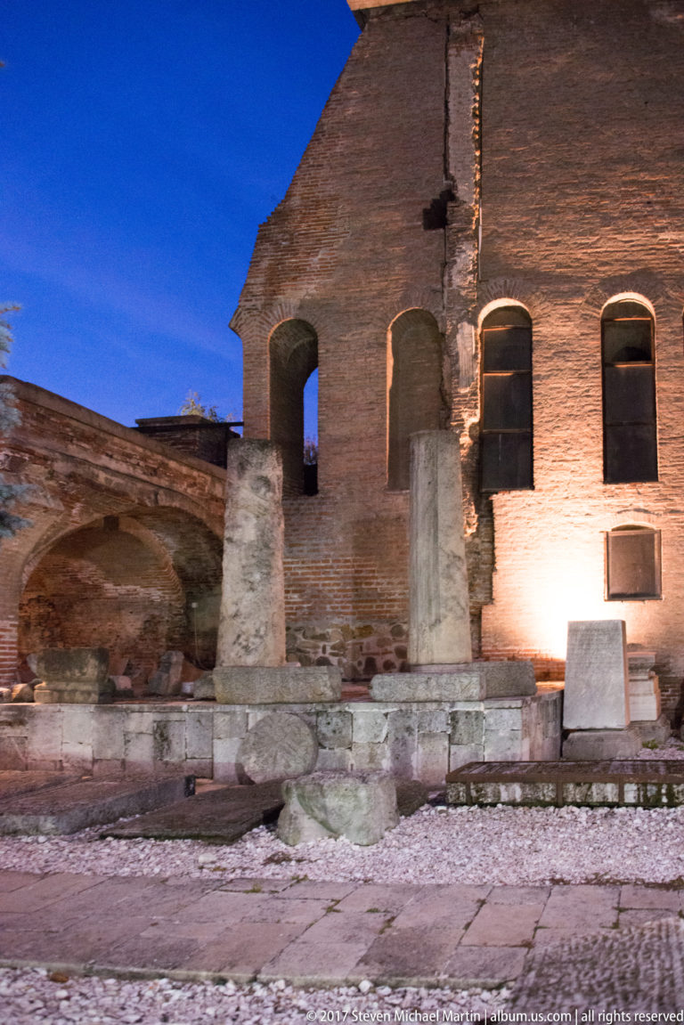 Night-lit ruined brick wall in Bucharest with tall arched openings, a major vertical crack, and stone fragments in front.