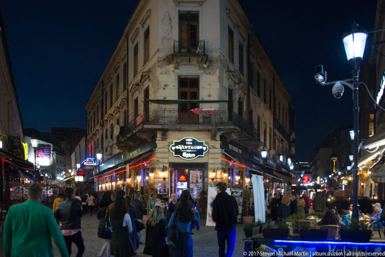 Crowded Bucharest nightlife corner with outdoor cafés beneath a weathered historic building and bright evening streetlights.