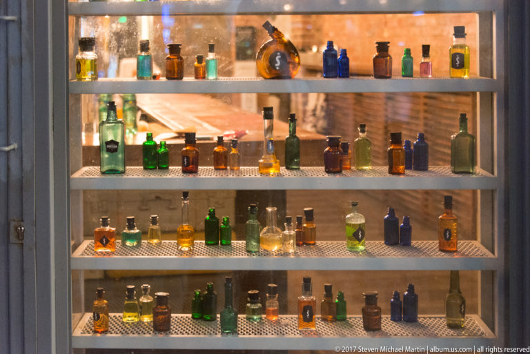 Backlit window display with four shelves of small green, amber, blue, and clear glass bottles in varied shapes.