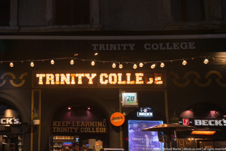Illuminated “TRINITY COLLEGE” bar sign with exposed bulbs and beverage branding above a dark Bucharest entrance.