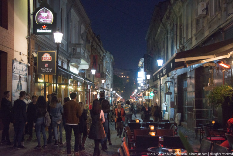 Narrow Bucharest pedestrian lane crowded at night, with restaurant terraces, awnings, hanging signs, lamps, and historic façades.