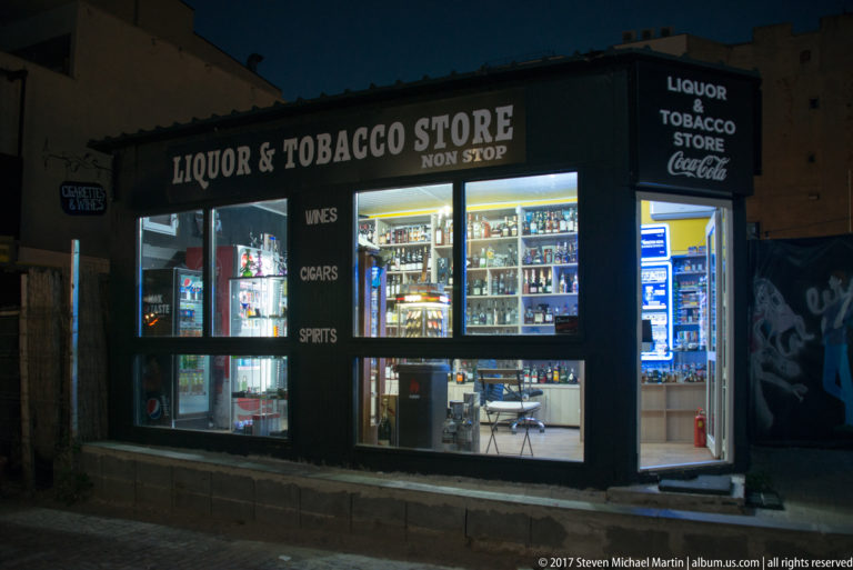 Bright Bucharest shop labeled “LIQUOR & TOBACCO STORE NON STOP,” with illuminated windows showing bottles and packaged goods.