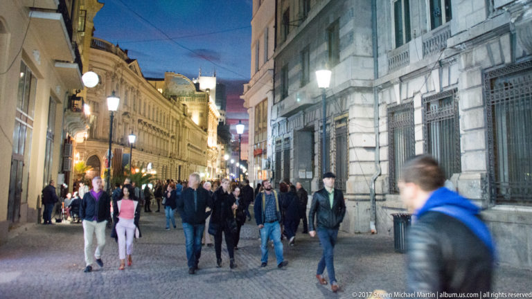 Pedestrians on a stone-paved Bucharest street at dusk between historic buildings, streetlamps, barred windows, and shopfronts.
