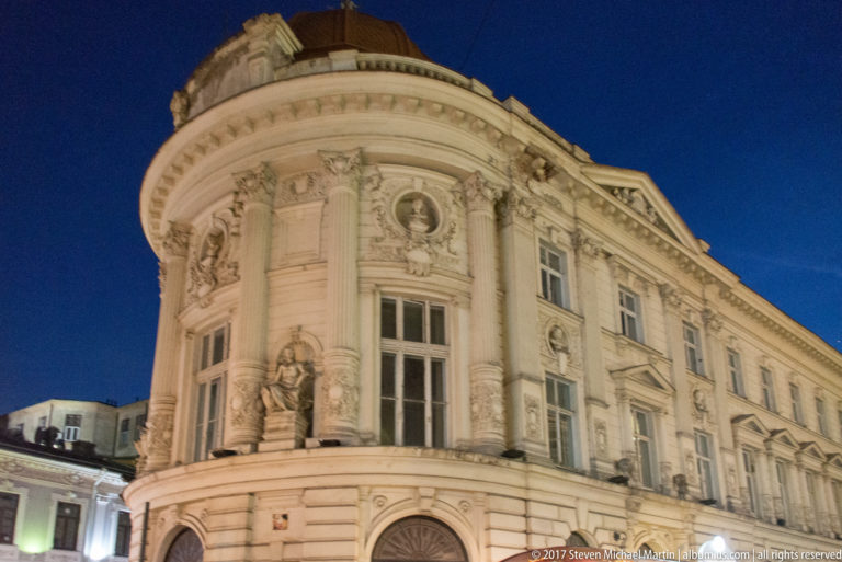 Illuminated rounded historic Bucharest façade with pilasters, sculpted figures, portrait medallions, floral ornament, and tall windows.