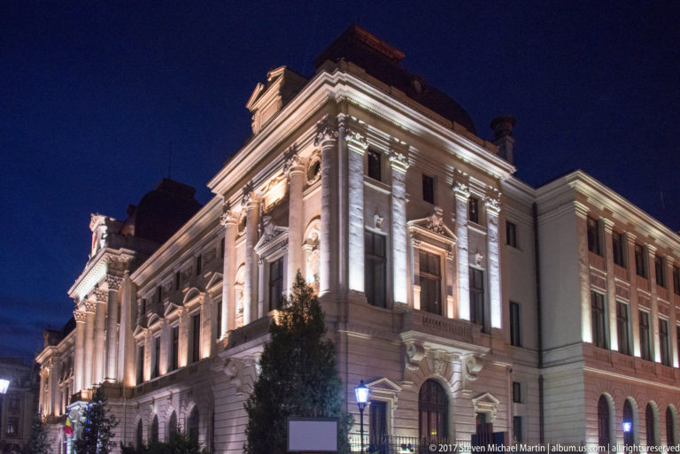 Grand illuminated Bucharest building with giant pilasters, tall windows, arched ground-floor openings, projecting bay, tree, and streetlamp.