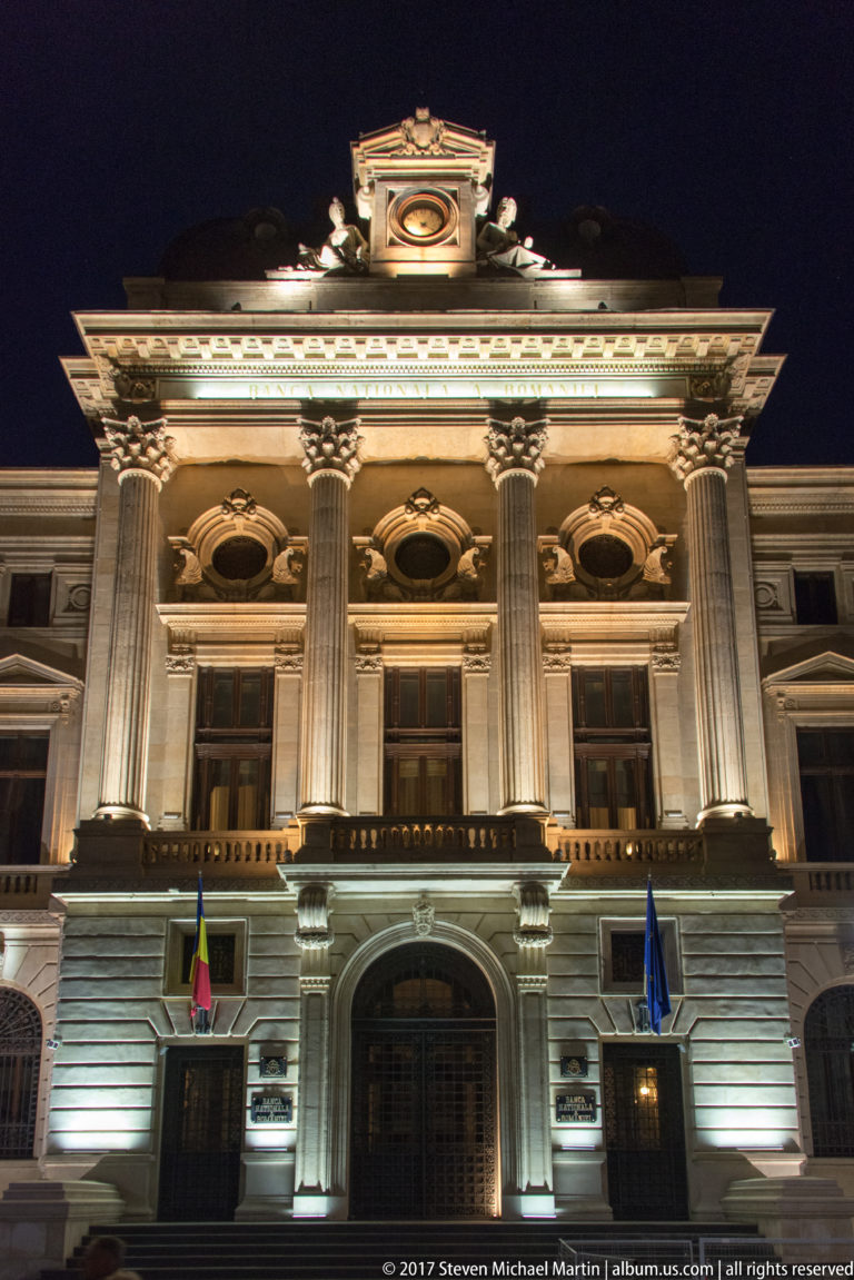 Illuminated National Bank of Romania façade with Corinthian columns, Romanian and EU flags, sculptural roofline, and central entrance at night.