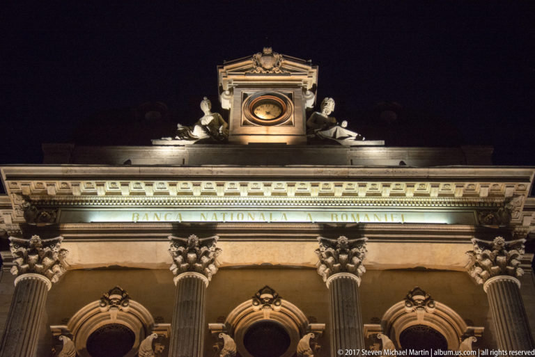 Low-angle National Bank of Romania façade with inscribed name, Corinthian capitals, arched windows, rooftop sculpture, and circular feature.