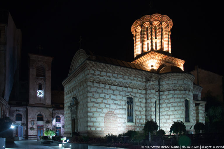 Small illuminated Orthodox church in Bucharest with striped brick-and-stone walls, decorative cornice, arched windows, and tall glowing tower.