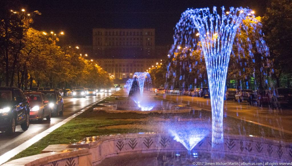 Blue illuminated fountains along a Bucharest boulevard median at night, with traffic, autumn trees, streetlights, and a large building beyond.