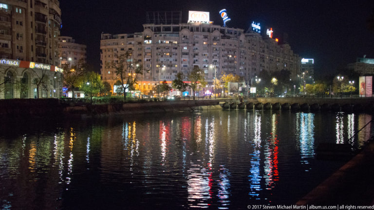 Bucharest buildings and rooftop signs at night reflected in dark water, with illuminated shops, streetlights, and trees along the opposite bank.