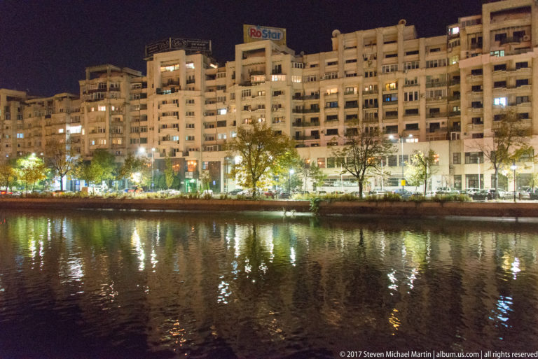 Long Bucharest apartment block with many balconies, illuminated windows, RoStar rooftop sign, trees, and reflections across water at night.
