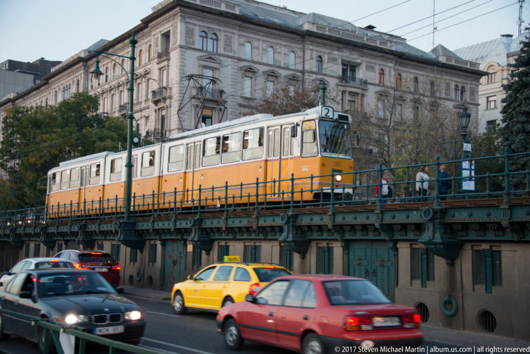 Yellow Budapest tram on a separated track with cars and taxis moving below beside a historic building.