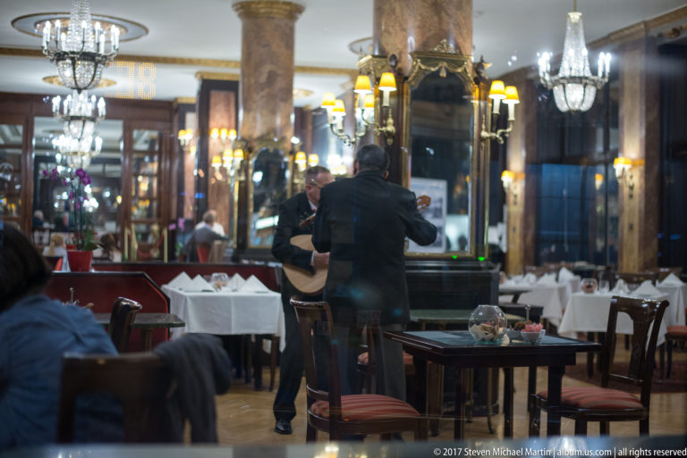 Elegant restaurant dining room with mirrors, marbleized columns, chandeliers, white tablecloths, diners, and staff.
