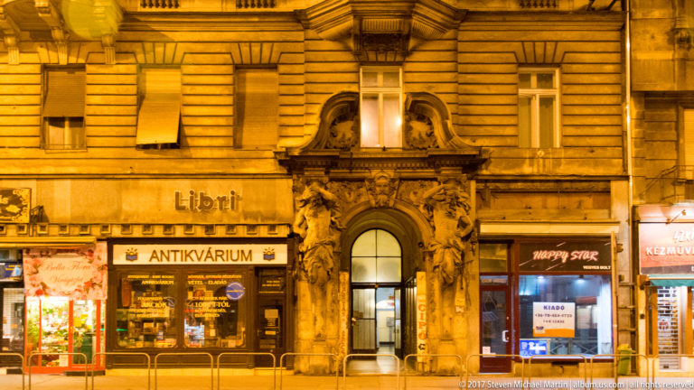 Illuminated historic façade with sculpted arched doorway and Libri and Antikvárium shop signs at street level.