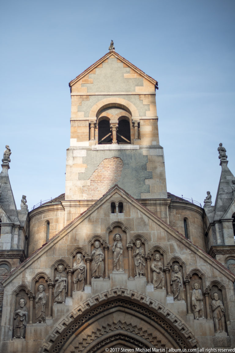 Romanesque Revival chapel façade with carved standing figures beneath the gable and a bell opening above.
