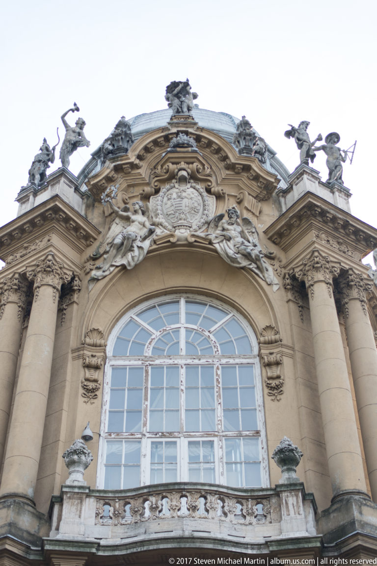 Large arched window framed by columns and crowned with a shield, scrollwork, and rooftop sculptures.