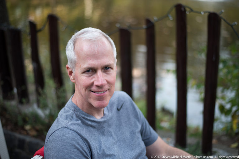 Gray-haired man smiling outdoors with tree trunks and softly blurred water behind him.