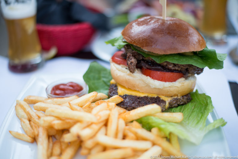 Double-patty hamburger with lettuce, tomato, cheese, sauce, and a pile of French fries on a white plate.