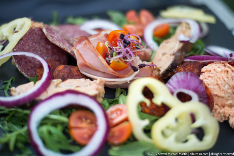 Close plate of sliced ham and salami with tomatoes, onion rings, peppers, greens, sprouts, and a seasoned spread.