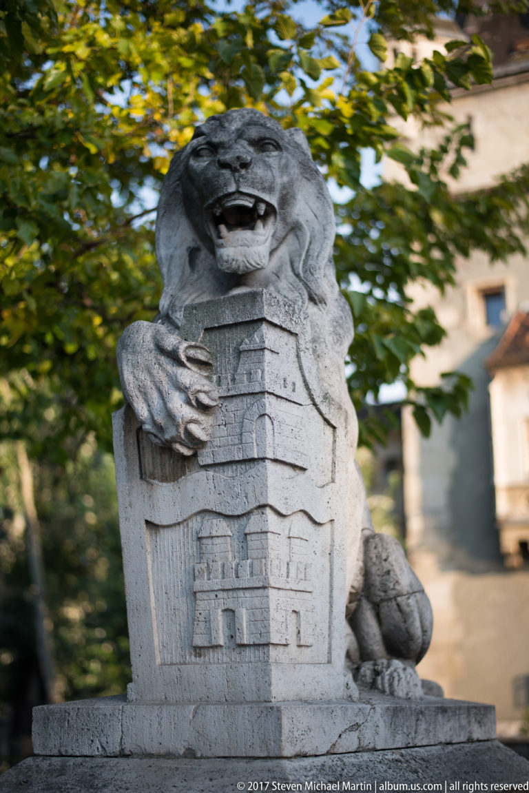 Weathered stone lion with open mouth and paw resting on a shield carved with a building façade.