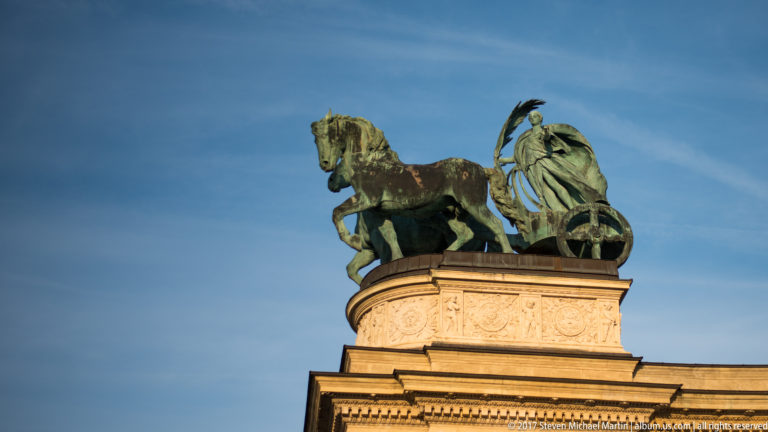 Green-patinated chariot sculpture with two horses and a standing female figure atop Heroes’ Square colonnade.