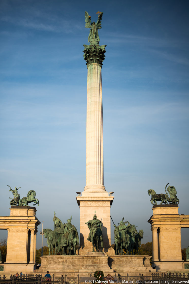 Full view of Heroes’ Square Millennium Monument with tall central column, equestrian statues, curved colonnades, and rooftop chariots.