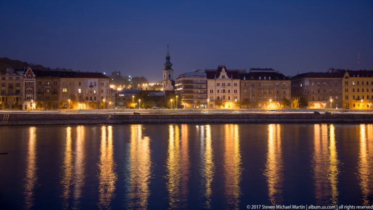 Night view across the Danube toward Buda buildings and a church spire, with golden reflections on the water.