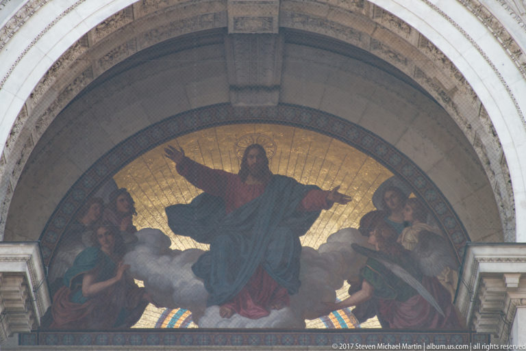 Gold-ground lunette mosaic of Christ with outstretched arms and angels above a basilica entrance.