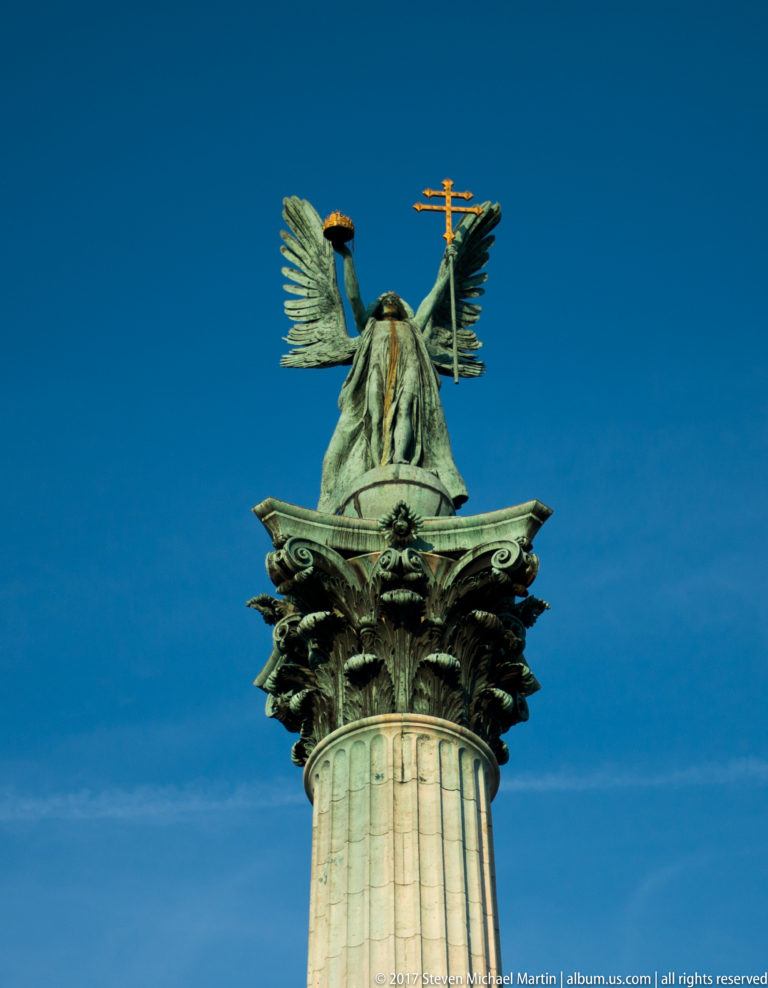 Green-patinated Archangel Gabriel with spread wings holding the Holy Crown and double cross atop a tall column.
