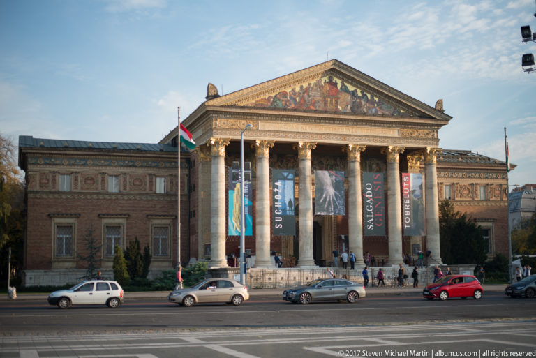 Frontal view of Budapest’s Műcsarnok with tall Corinthian columns, decorated pediment, exhibition banners, and cars in front.