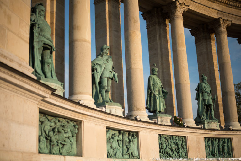 Bronze historical figures standing between stone columns above rectangular relief panels in the Heroes’ Square colonnade.