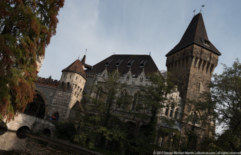 Tilted moat-side view of Vajdahunyad Castle with square tower, conical turret, steep roofs, and trees.