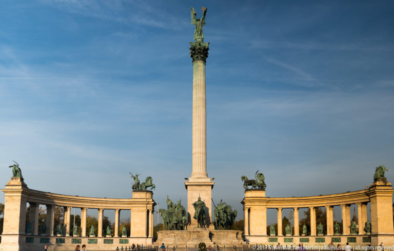 Wide frontal view of Heroes’ Square with Millennium Column, equestrian statues, curved colonnades, and rooftop chariots.