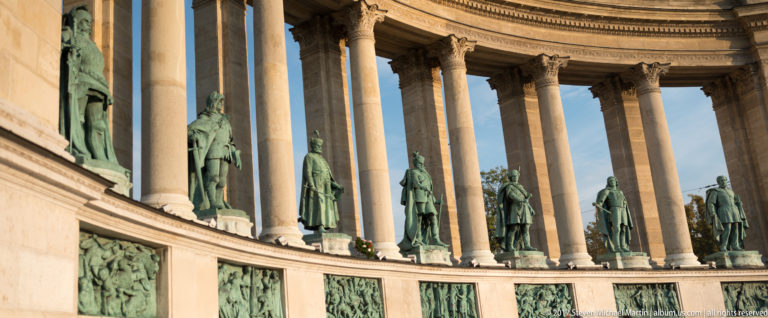 Oblique view along Heroes’ Square colonnade with bronze historical figures between columns and relief panels below.