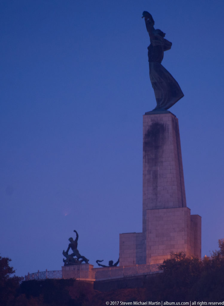 Silhouetted Liberty Statue with palm-bearing female figure and smaller bronze groups against a deep blue evening sky.
