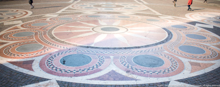 Large circular geometric paving design in pink, gray, white, and blue-gray stone across St. Stephen’s Square.