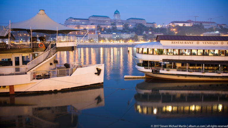 White excursion and restaurant boats moored on the Danube with illuminated Buda Castle reflected across the water.