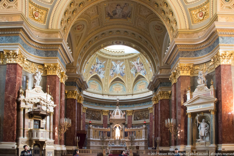 Wide sanctuary view with red marble piers, painted apse, gilded capitals, side monuments, and central altar.